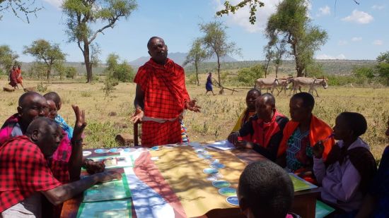 Foto de miembros de la tribu Maasai reunidos alrededor de una mesa, charlando y mirando los materiales del taller.